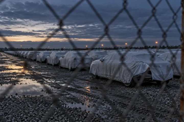 Aire d'entreposage de voitures neuves sous housses blanches, derrière une clôture grillagée, au crépuscule