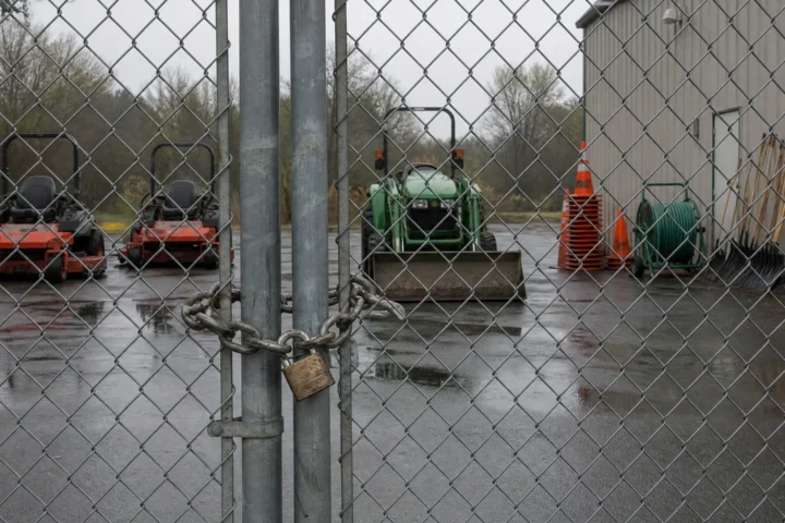 Cour d'entretien municipale fermée par une clôture cadenassée, tondeuses et outils immobiles derrière la grille.