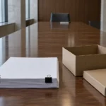 Une épaisse liasse de documents et une boîte d’archives au bout d’une longue table de salle d’audience, face à une chaise vide.