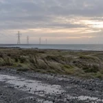 Terrain industriel côtier au petit jour, pylônes et éoliennes lointaines dans la brume