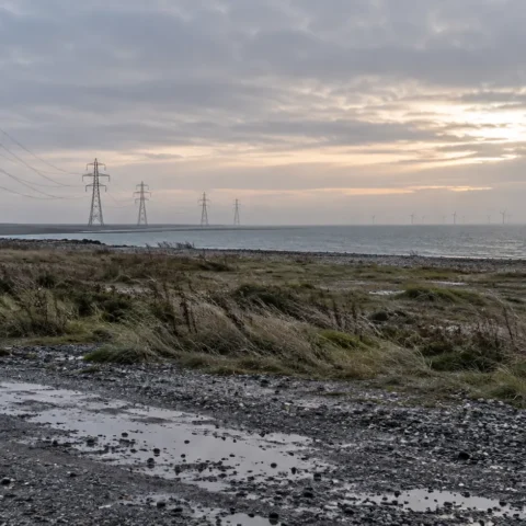 Terrain industriel côtier au petit jour, pylônes et éoliennes lointaines dans la brume