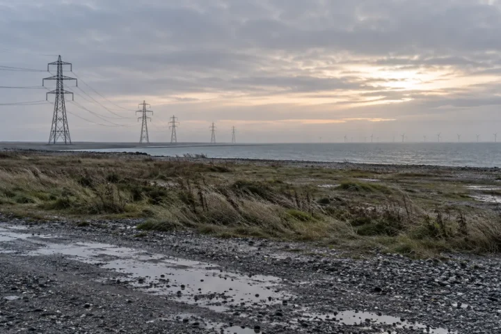 Terrain industriel côtier au petit jour, pylônes et éoliennes lointaines dans la brume