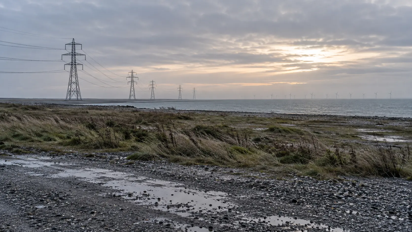 Terrain industriel côtier au petit jour, pylônes et éoliennes lointaines dans la brume