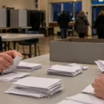 Mains de scrutateurs triant à la main des bulletins de vote près d'une urne sans emblème, dans un bureau de vote en fin de soirée